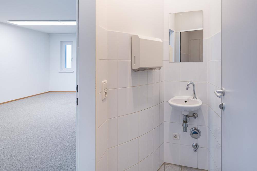 Kleiner Waschbereich mit Waschbecken, Spiegel und Seifenspender, angrenzend an einen Büroraum.