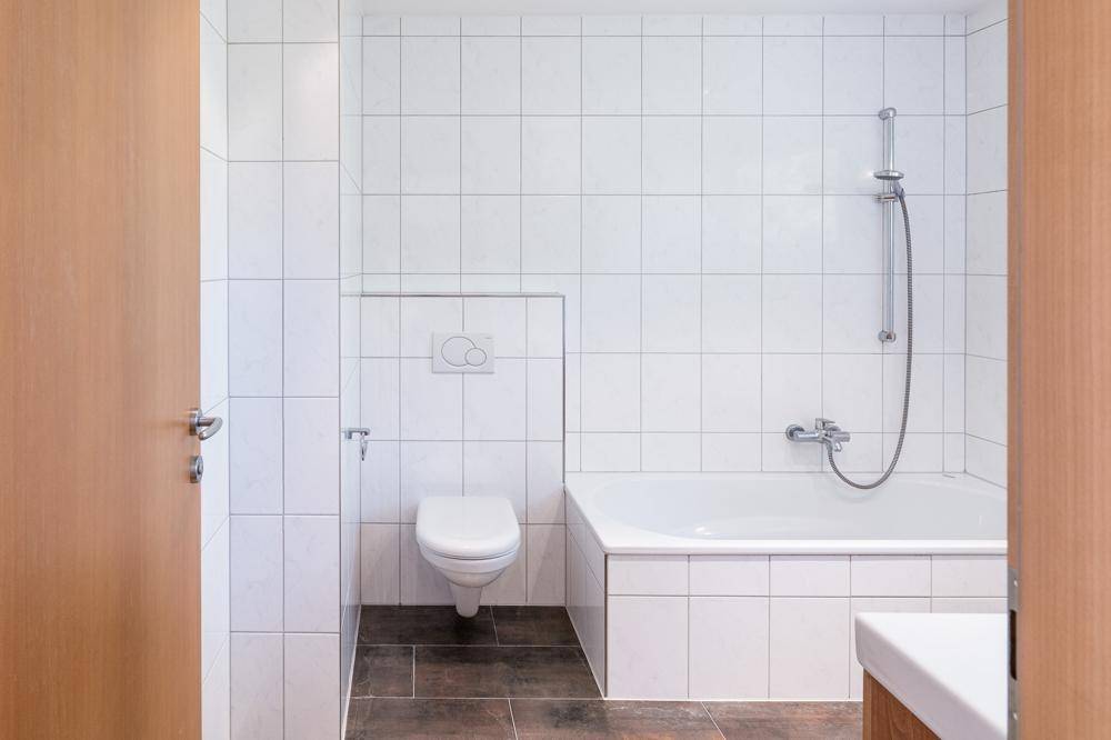 Badezimmer mit Badewanne, Toilette und weißen Fliesen, durch eine Holztür zugänglich.