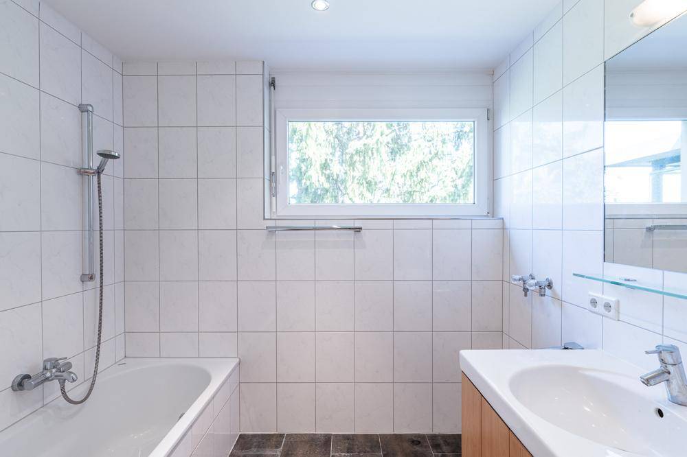 Modernes Badezimmer mit Badewanne, Waschbecken, Fenster und weißen Wandfliesen.