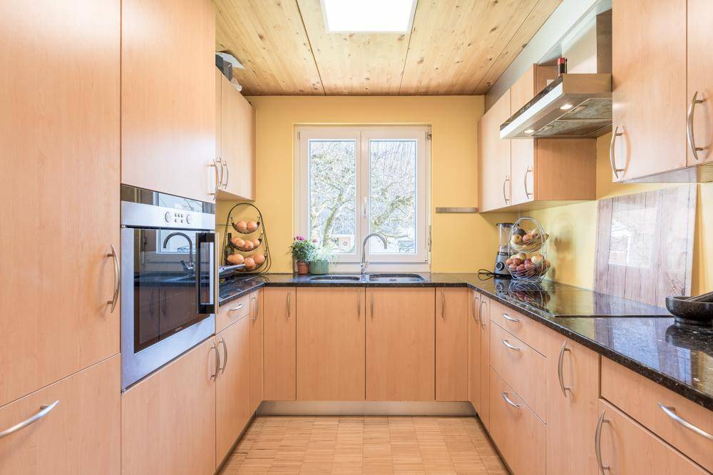 Funktionale Küche mit hellen Holzschränken, dunkler Arbeitsplatte und Fenster mit Blick ins Grüne.