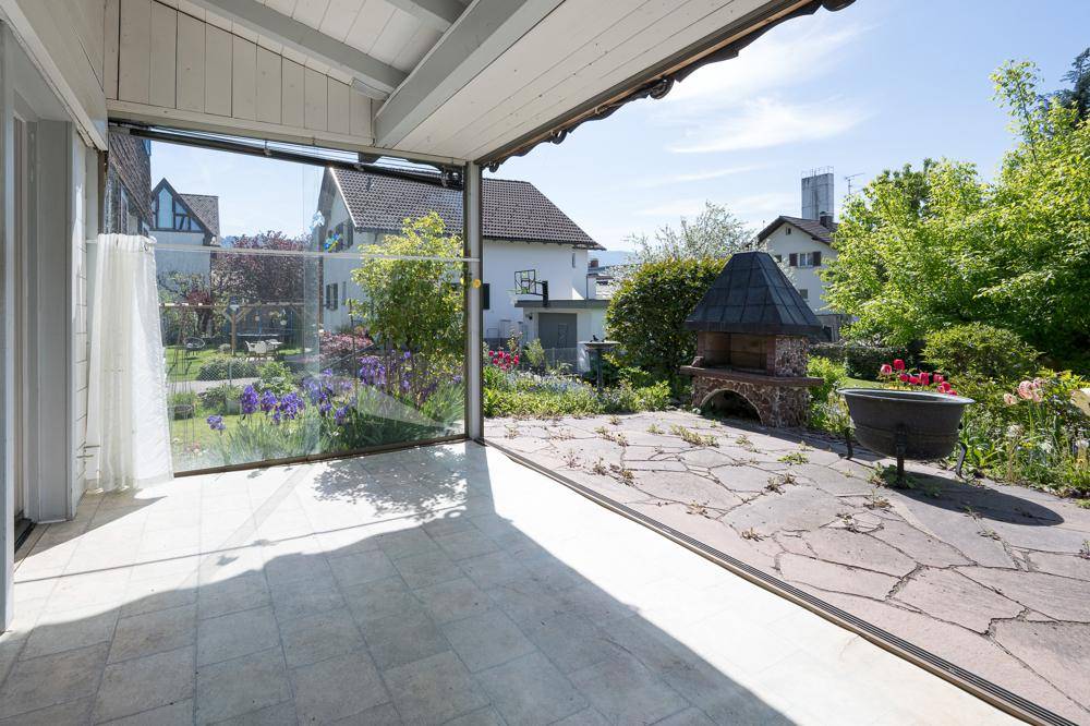 Verglaster Wintergarten mit Zugang zu einer gepflasterten Terrasse und Grillplatz im Garten.