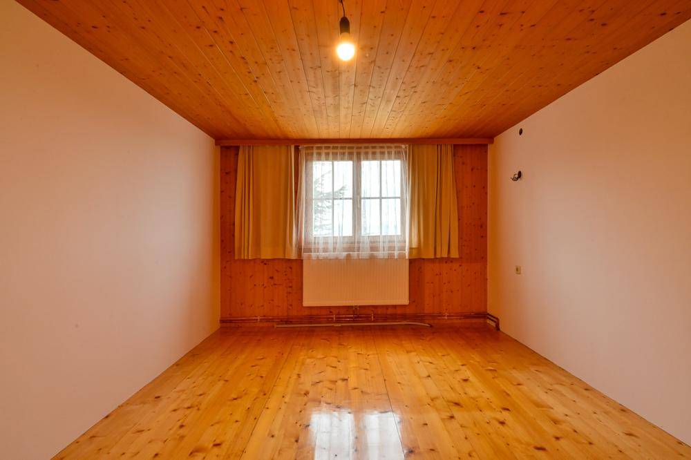 Helles Zimmer mit Holzdecken, Holzboden und einer großen Fensterfront mit Vorhängen.