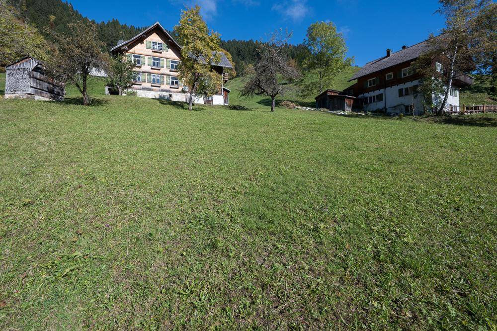 Grüne Wiese mit traditionellen Häusern am Hang und einem weiten Blick auf die umliegende Berglandschaft.