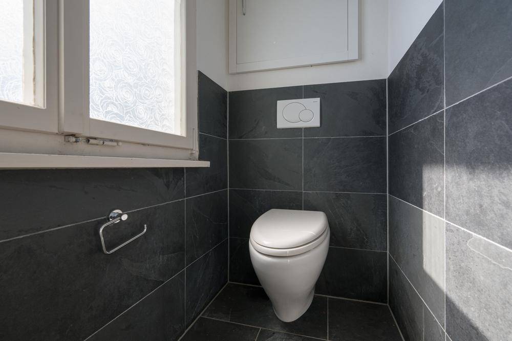 Separate Toilette mit Wand-WC, dunklen Fliesen und einem Fenster.