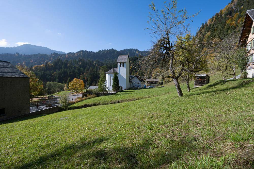 Panoramablick über eine grüne Wiese mit einer Kirche und bewaldeten Bergen im Hintergrund an einem sonnigen Tag.