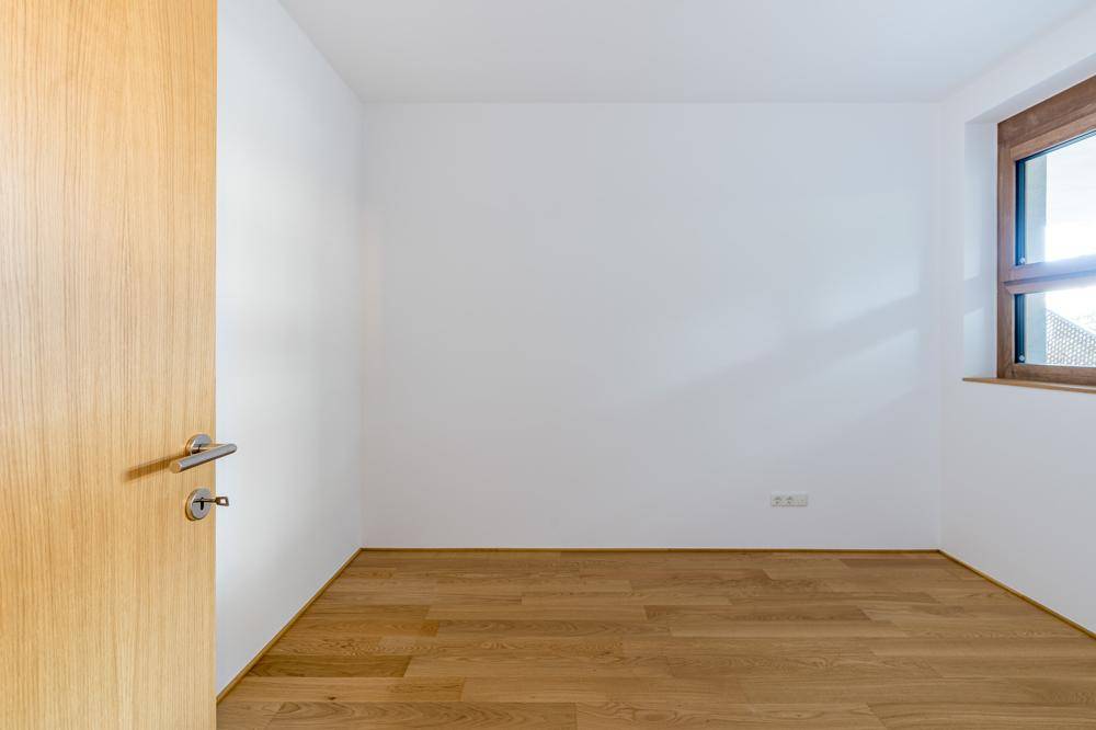 Helles, leeres Zimmer mit Holzboden und einem Fenster mit Blick ins Grüne.