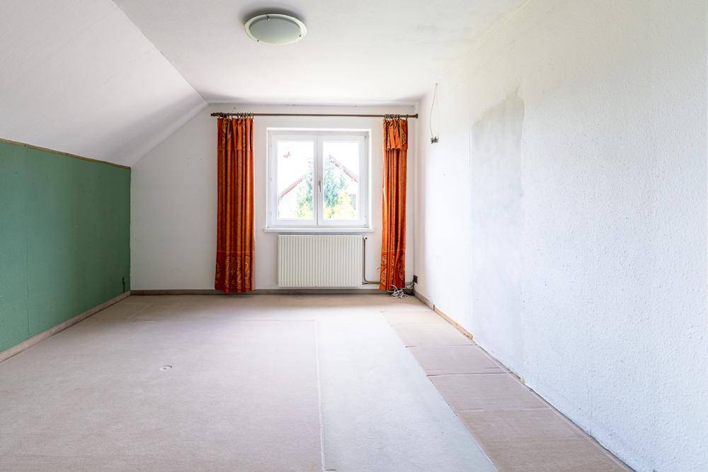 Dachzimmer mit schrägen Wänden, Teppichboden und einem Fenster mit Blick ins Grüne.