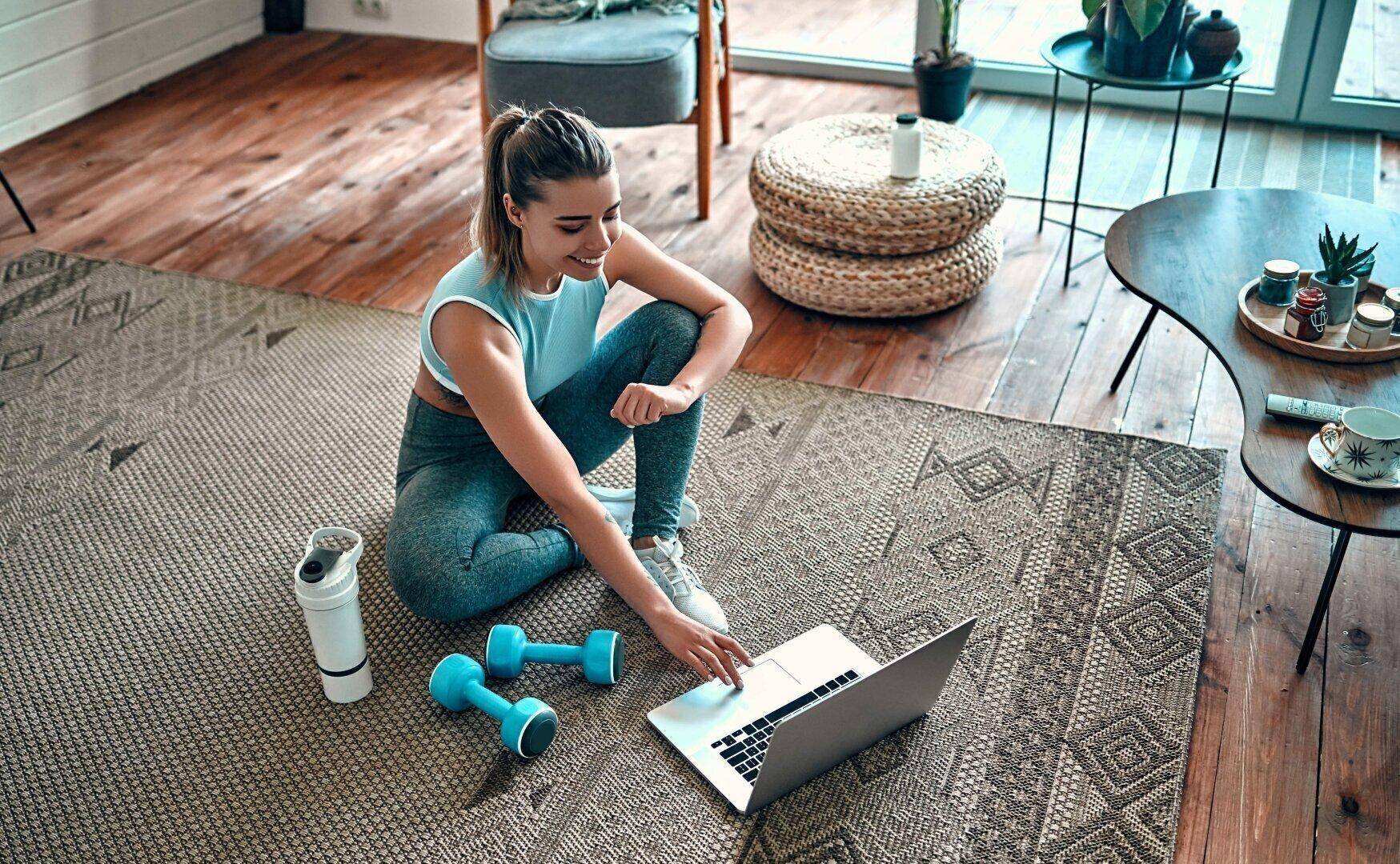 Gemütliches Wohnzimmer mit Holzboden und Teppich, genutzt als Home-Gym mit Hanteln und Laptop.