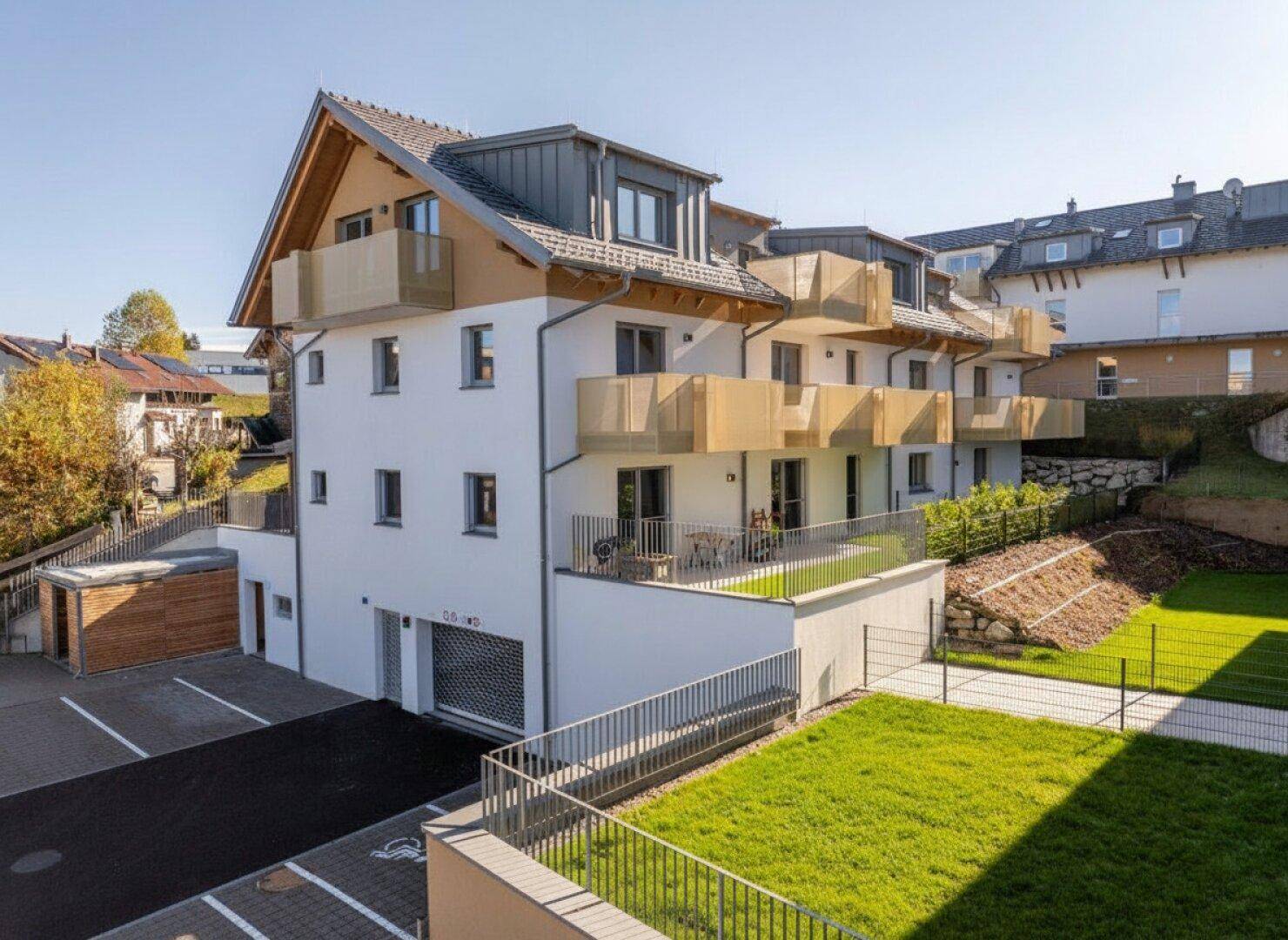 Modernes Mehrfamilienhaus mit Balkonen, Grünflächen und Außenparkplätzen an einem sonnigen Tag.
