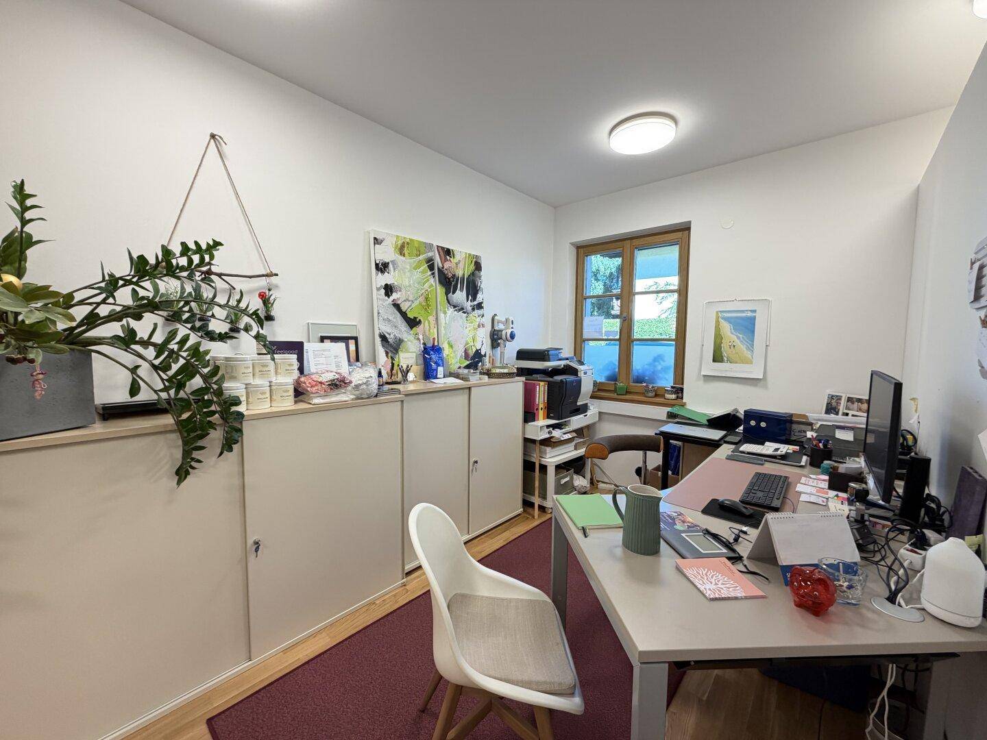 Ein aufgeräumtes Büro mit einem Schreibtisch, Stühlen, Schränken und einem Fenster mit Blick ins Grüne.