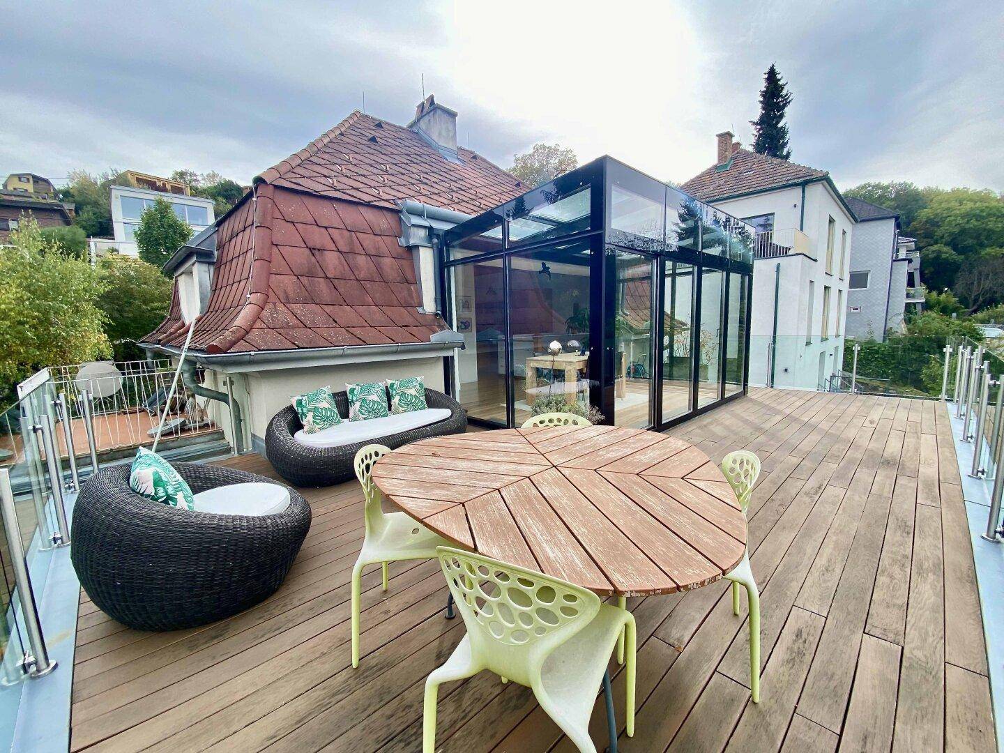 Große Holzterrasse mit Esstisch und Lounge-Bereich, angrenzend an einen Wintergarten.