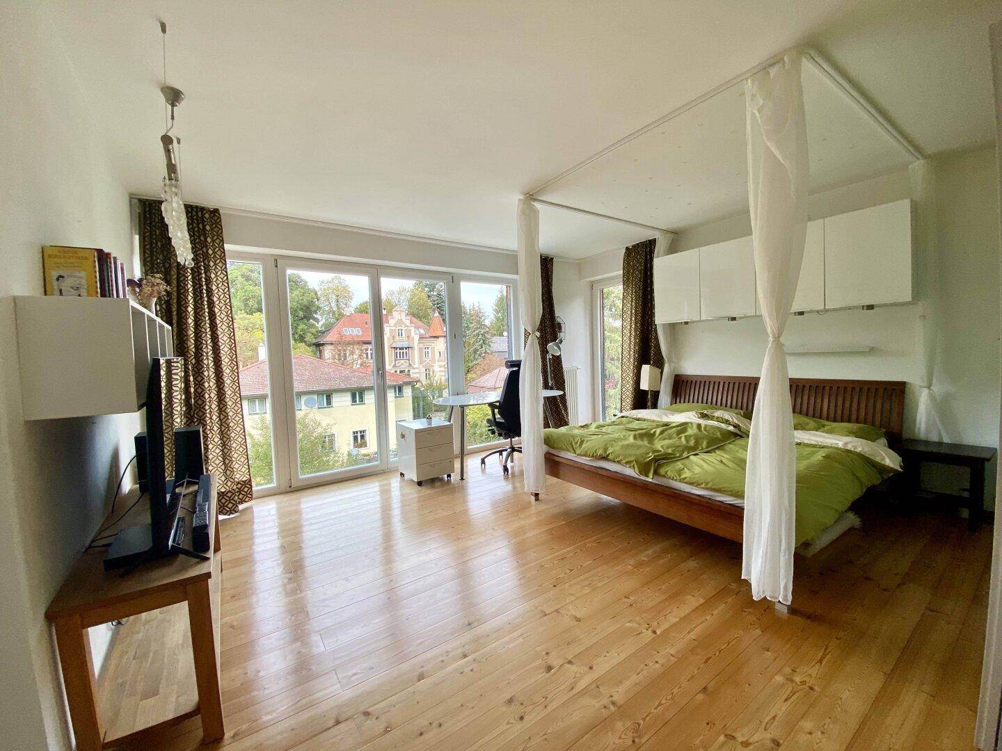 Geräumiges Schlafzimmer mit Holzboden, großen Fenstern und Blick ins Grüne.