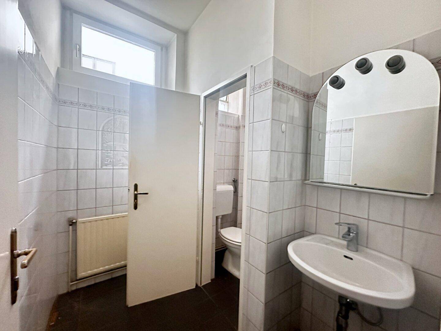 Gefliestes Badezimmer mit Waschbecken, Spiegel, Toilette und einem Fenster für Tageslicht.