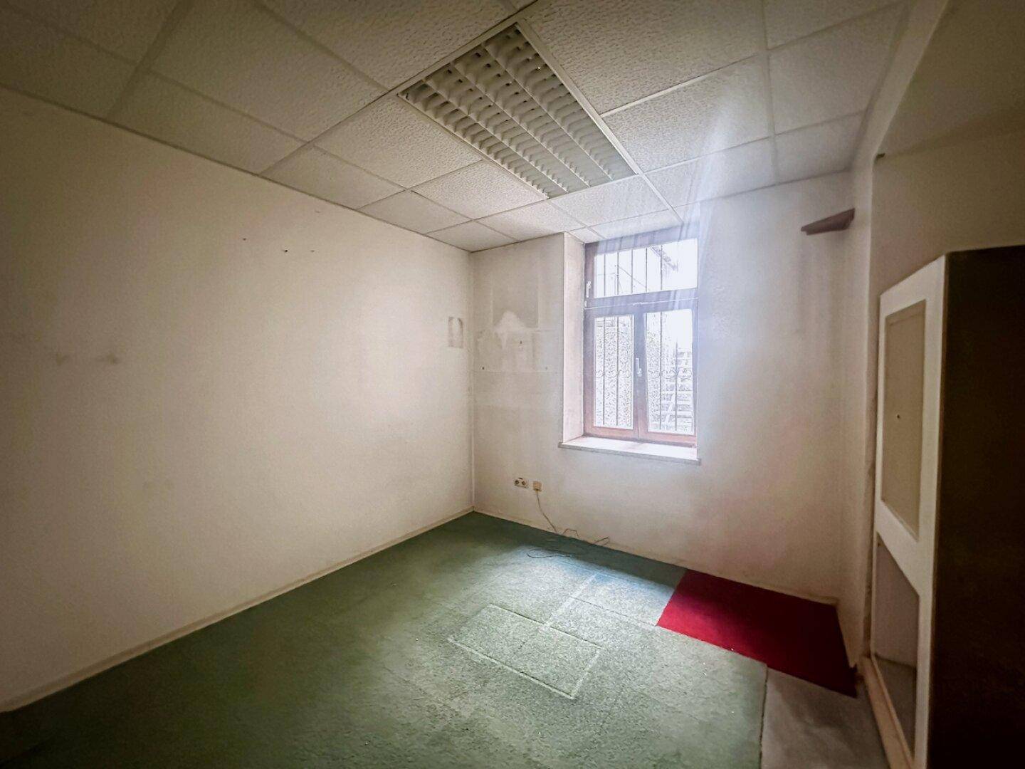 Kleiner Büroraum mit grünem Teppichboden und einem Fenster mit Gitter, ideal für ruhige Arbeit.
