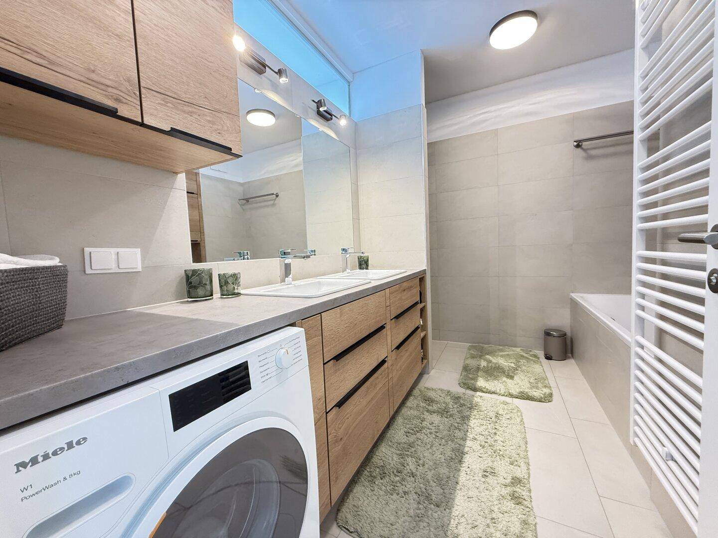 Modernes Badezimmer mit Doppelwaschbecken, Badewanne und integrierter Waschmaschine von Miele.