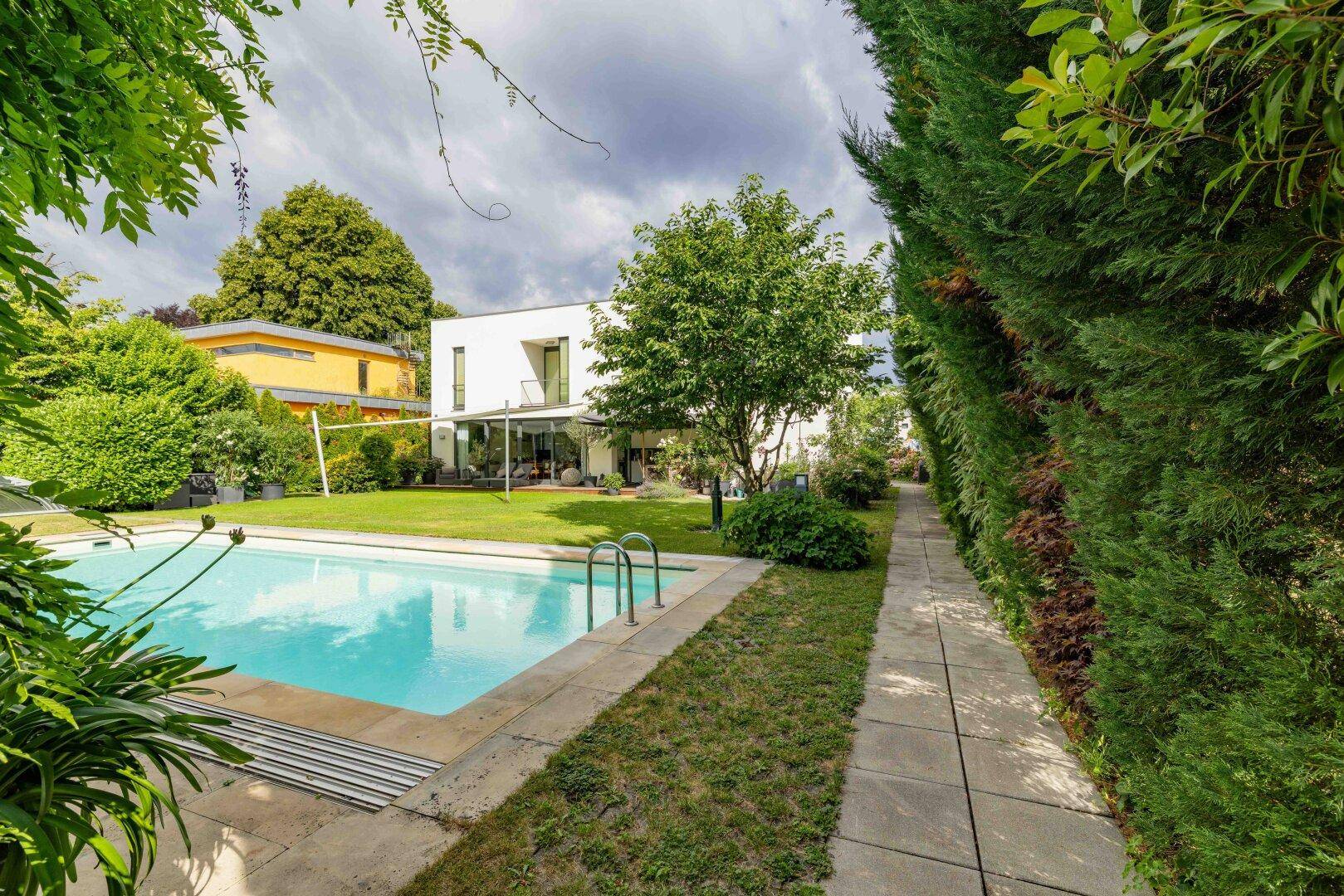 Großer Außenpool im Garten mit umgebender Hecke und Blick auf das moderne Wohnhaus.