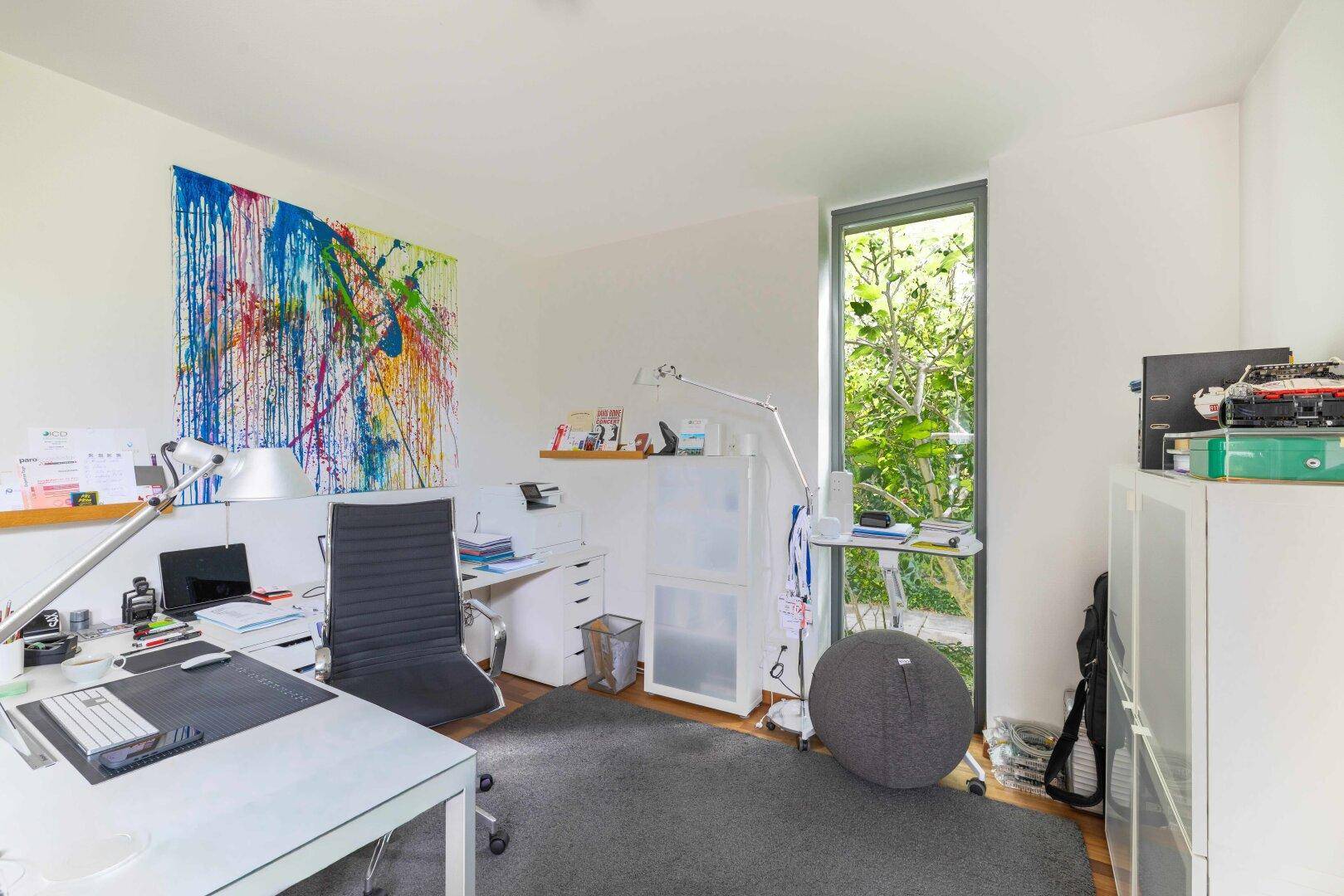 Helles Arbeitszimmer mit Schreibtisch, farbenfroher Kunst und großem Fenster zum Garten.