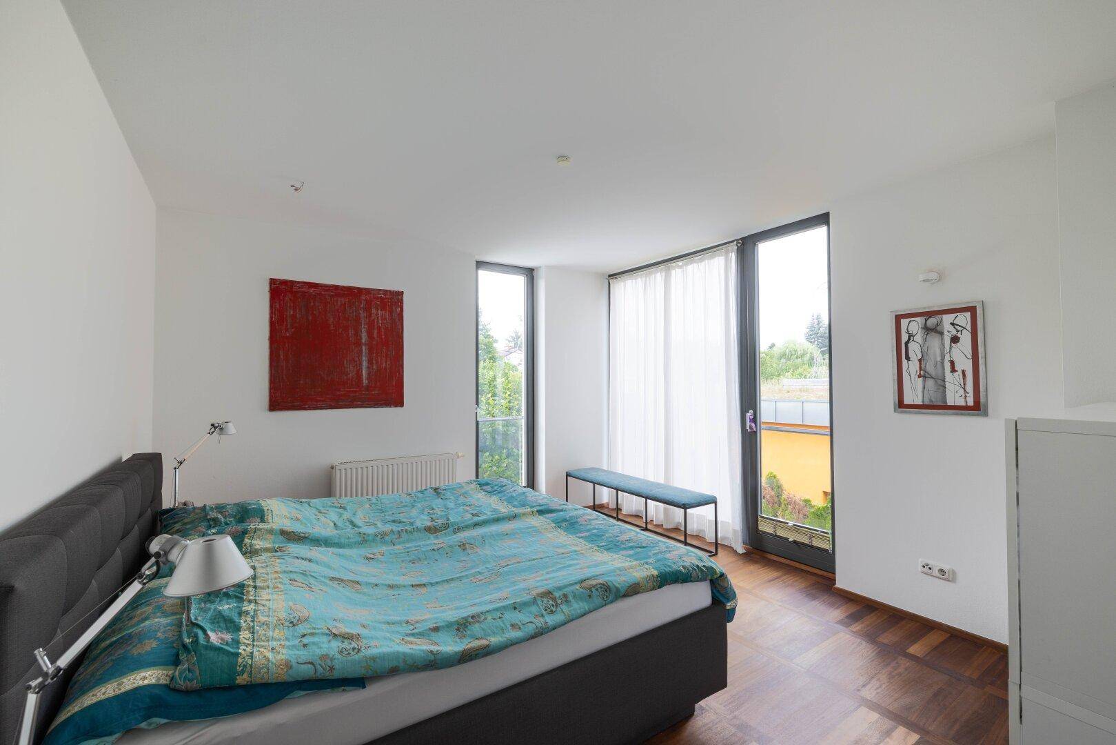 Helles Schlafzimmer mit Doppelbett, Holzboden und großen Fenstern mit Blick ins Grüne.