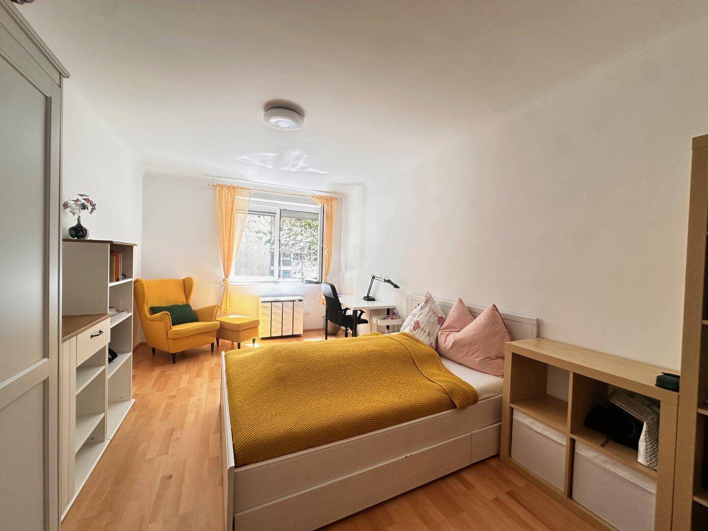 Helles Schlafzimmer mit Holzboden, Einzelbett, Schreibtisch und einem farbenfrohen Sessel am Fenster.