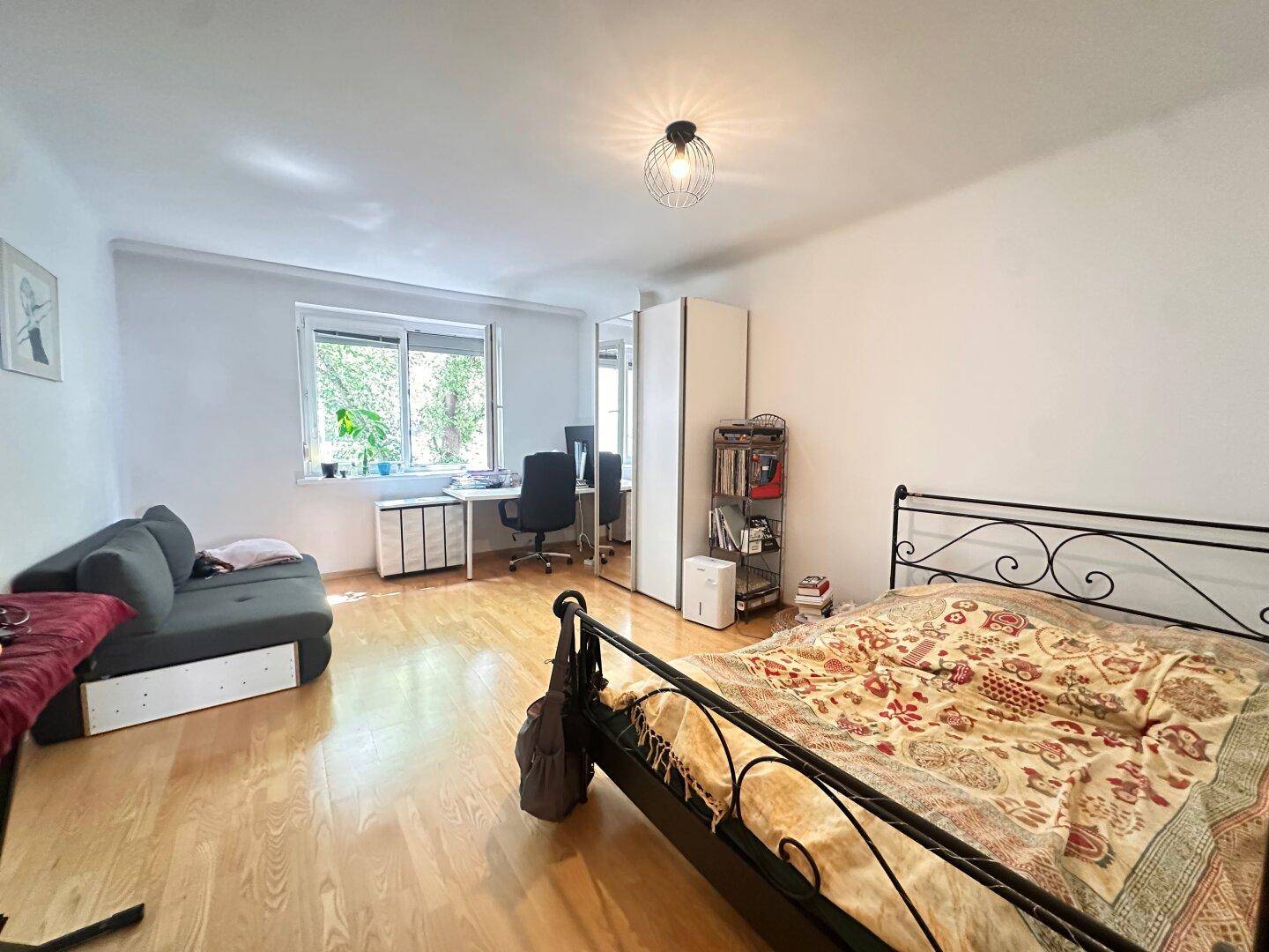 Geräumiges Schlafzimmer mit Holzboden, Metallbett, Sofa und einem Arbeitsbereich am Fenster.