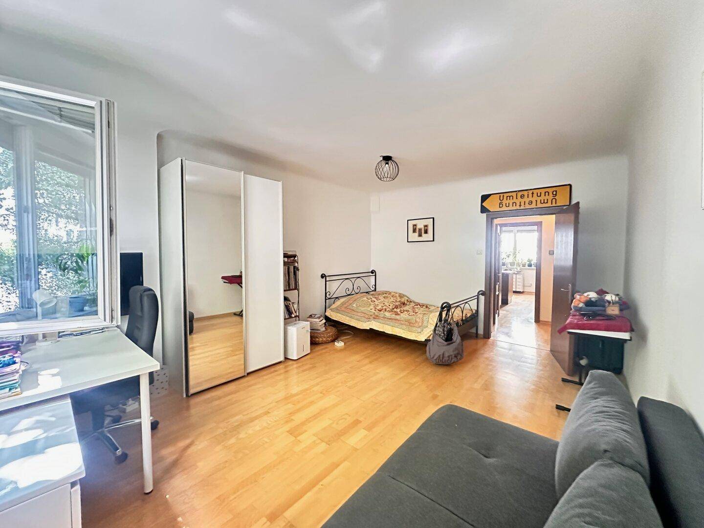 Helles Schlafzimmer mit Holzboden, Sofa, Bett und einem Durchgang zu einem weiteren Raum.