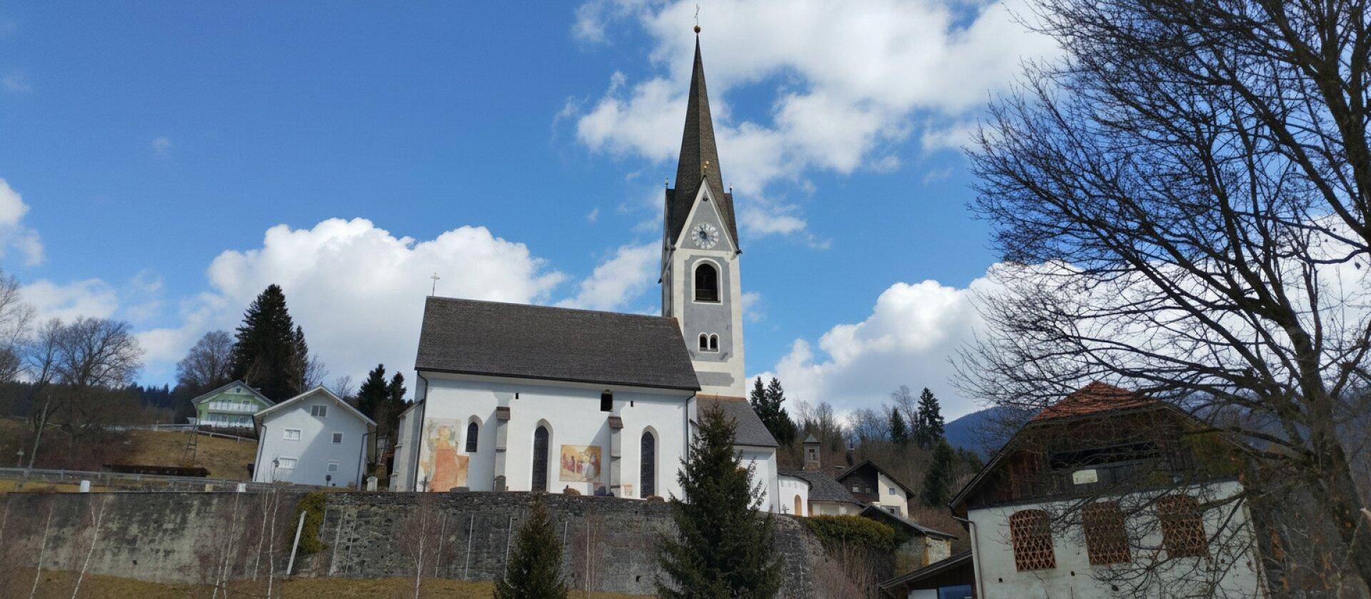 Historische weiße Kirche mit hohem Turm auf einem Hügel, umgeben von Bäumen und Häusern.