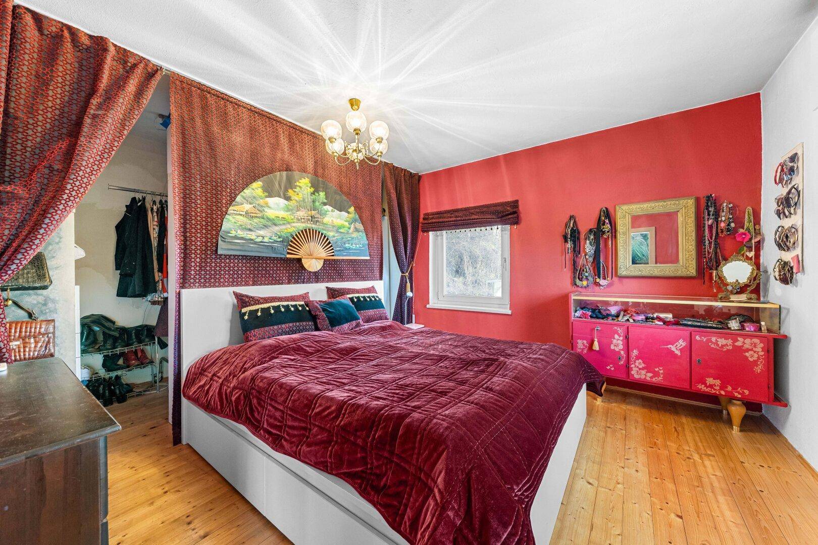 Schlafzimmer mit roter Wand, großem Bett und Vorhängen, die einen begehbaren Kleiderschrank abtrennen.