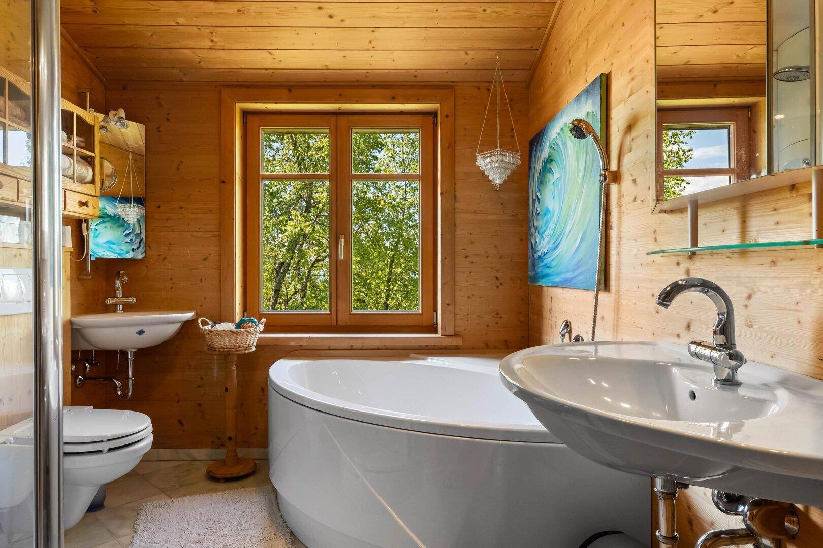 Helles Badezimmer mit Holzverkleidung, Badewanne, Dusche und Fenster mit Blick ins Grüne.