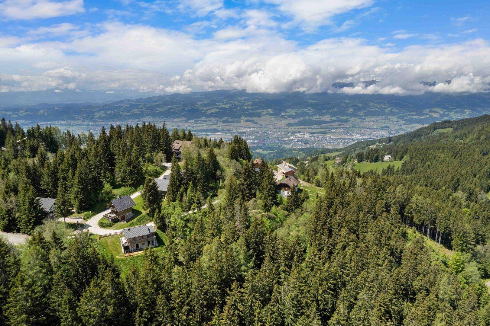 Weitläufige Luftaufnahme einer bewaldeten Berglandschaft mit Häusern und Fernblick ins Tal.