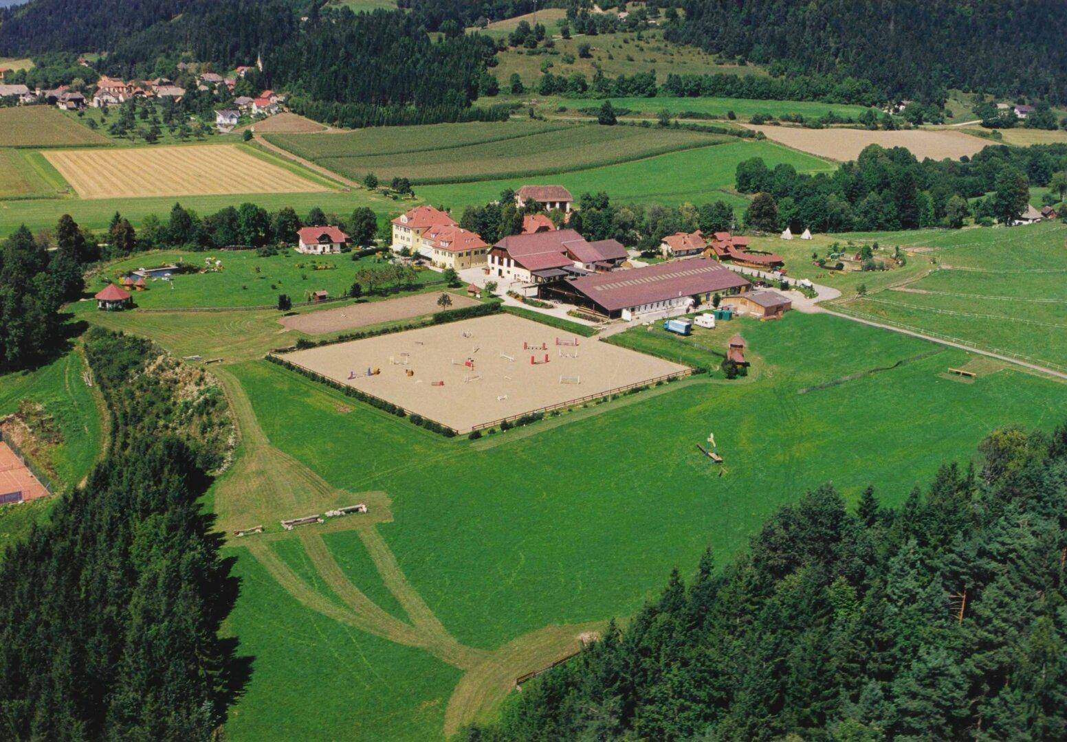 Weitläufiges Anwesen mit Gebäuden, Reitplatz und grünen Feldern, umgeben von Wäldern und Bergen.