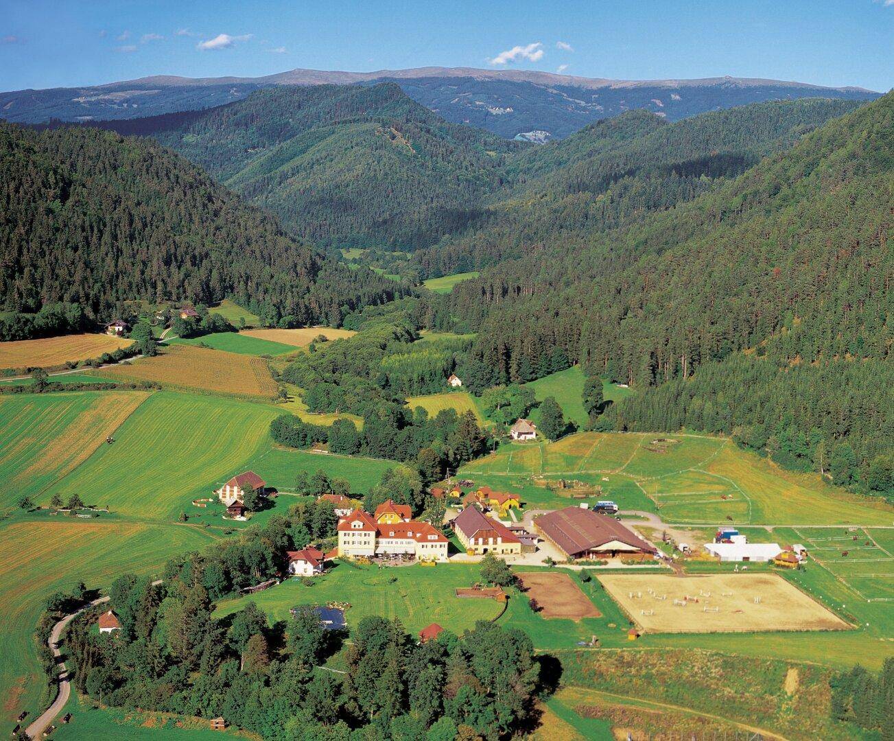 Großes Anwesen in einem grünen Tal, umgeben von Wäldern und Bergen, mit Reitplatz und Gebäuden.