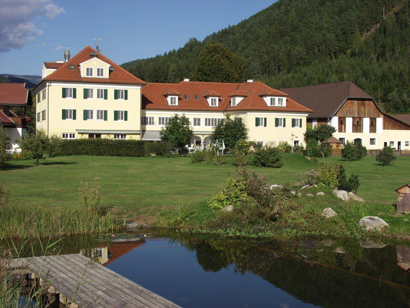 Großes Anwesen mit gelbem Gebäude und rotem Dach, umgeben von einem grünen Garten und einem Teich.