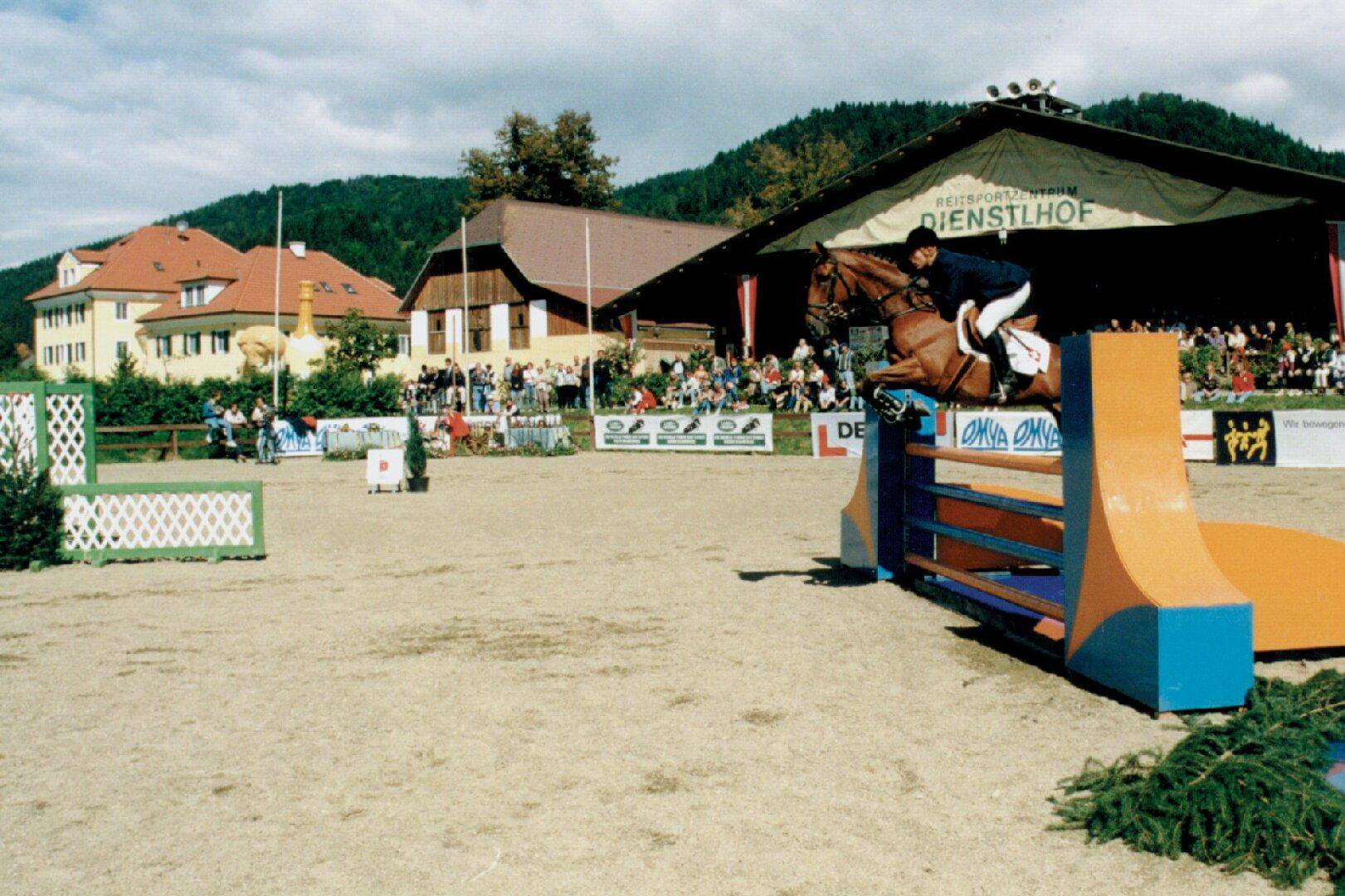 Pferdespringturnier auf dem Reitplatz mit Zuschauern und dem Hauptgebäude im Hintergrund.