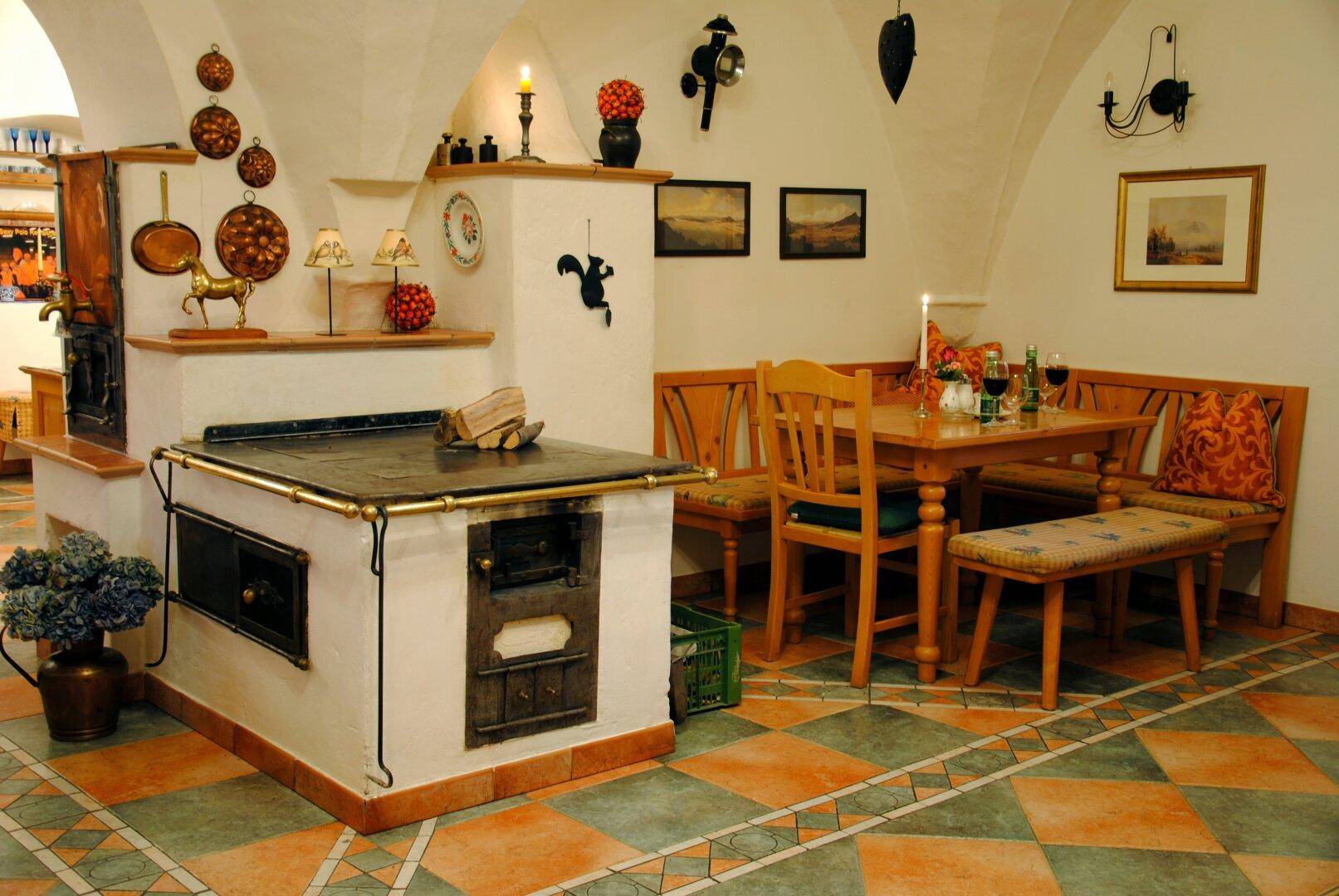 Rustikaler Essbereich mit traditionellem Kachelofen, Holzbänken und gemusterten Fliesenböden.