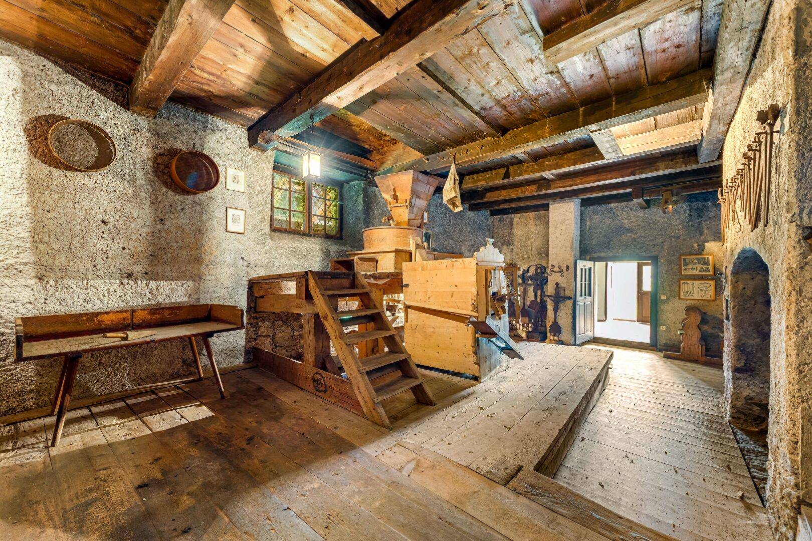 Historischer Raum mit Holzbalken, Steinwänden und alten Maschinen, möglicherweise eine Mühle.