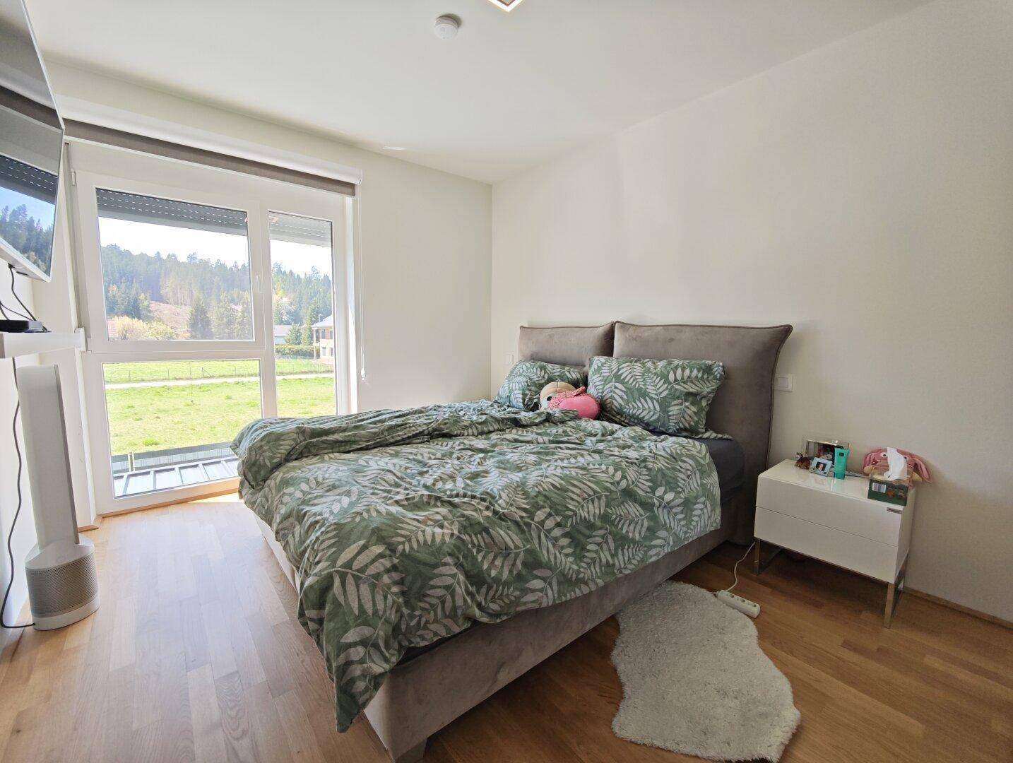 Helles Schlafzimmer mit großem Fenster, Holzboden und weitem Blick ins Grüne.