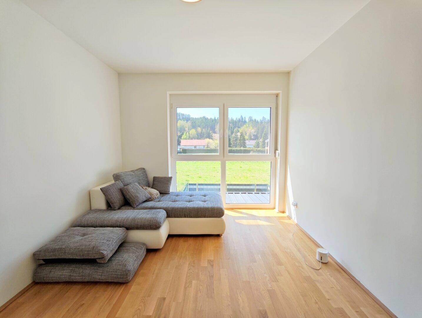 Freundliches Zimmer mit Sofa, Holzboden und großem Fenster mit Blick ins Grüne.