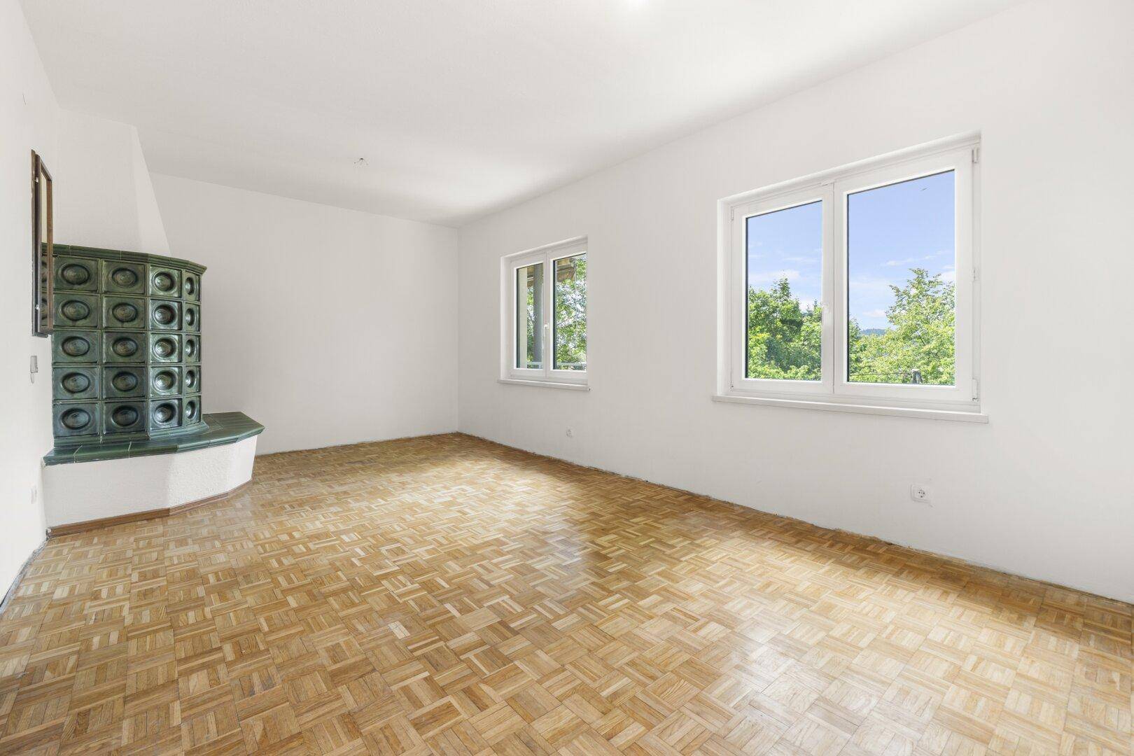 Helles Wohnzimmer mit Parkettboden, großem Fenster und einem traditionellen Kachelofen.