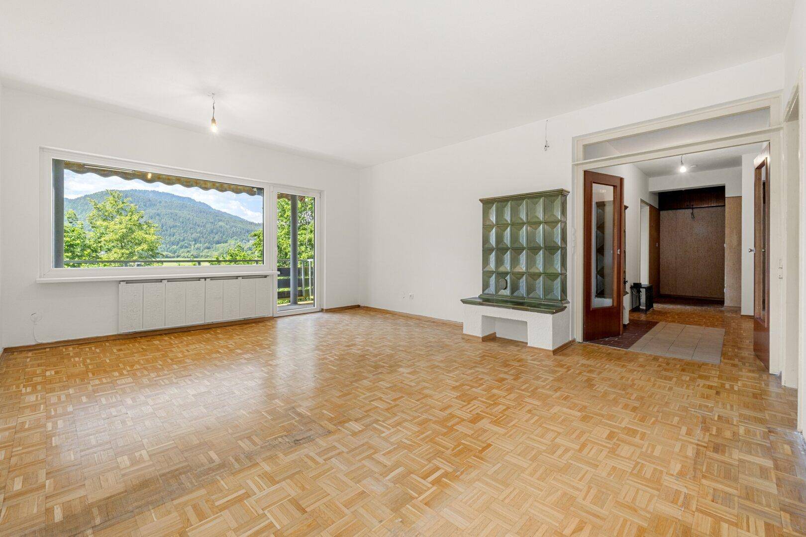 Lichtdurchflutetes Wohnzimmer mit Parkettboden, Kachelofen und Zugang zu einem Balkon mit Bergblick.