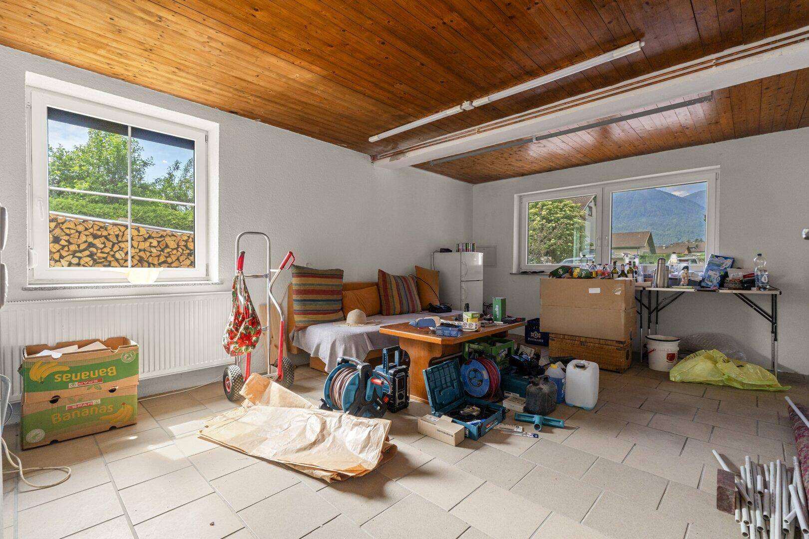 Vielseitiger Raum mit Holzdecke, Fliesenboden und Fenstern mit Blick ins Grüne und auf die Berge.