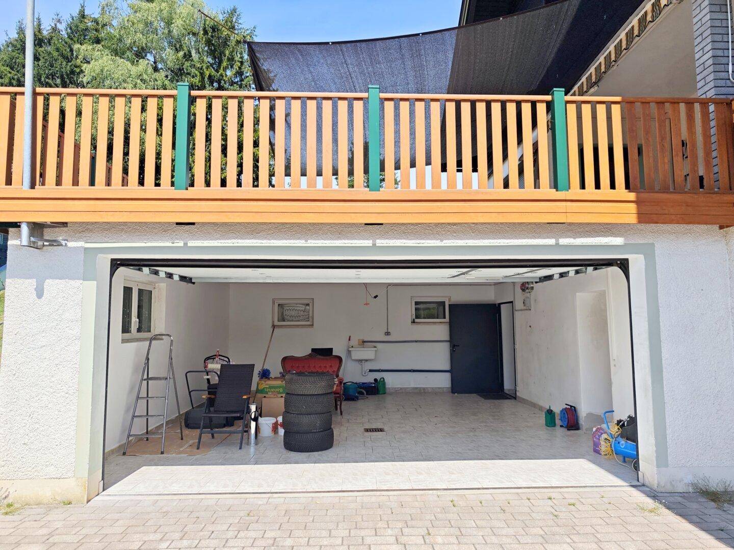 Offene Garage mit Fliesenboden und darüberliegendem Holzbalkon mit Geländer.