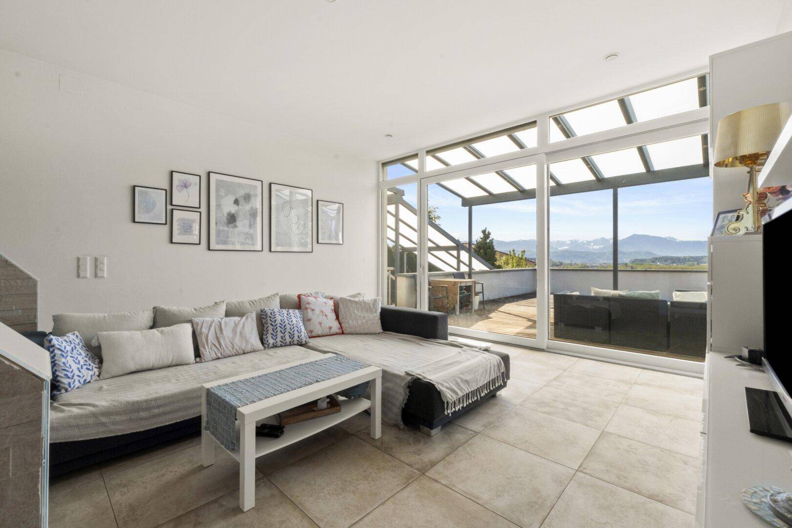 Geräumiges Wohnzimmer mit großem Sofa, Fliesenboden und Zugang zur überdachten Terrasse mit Bergblick.