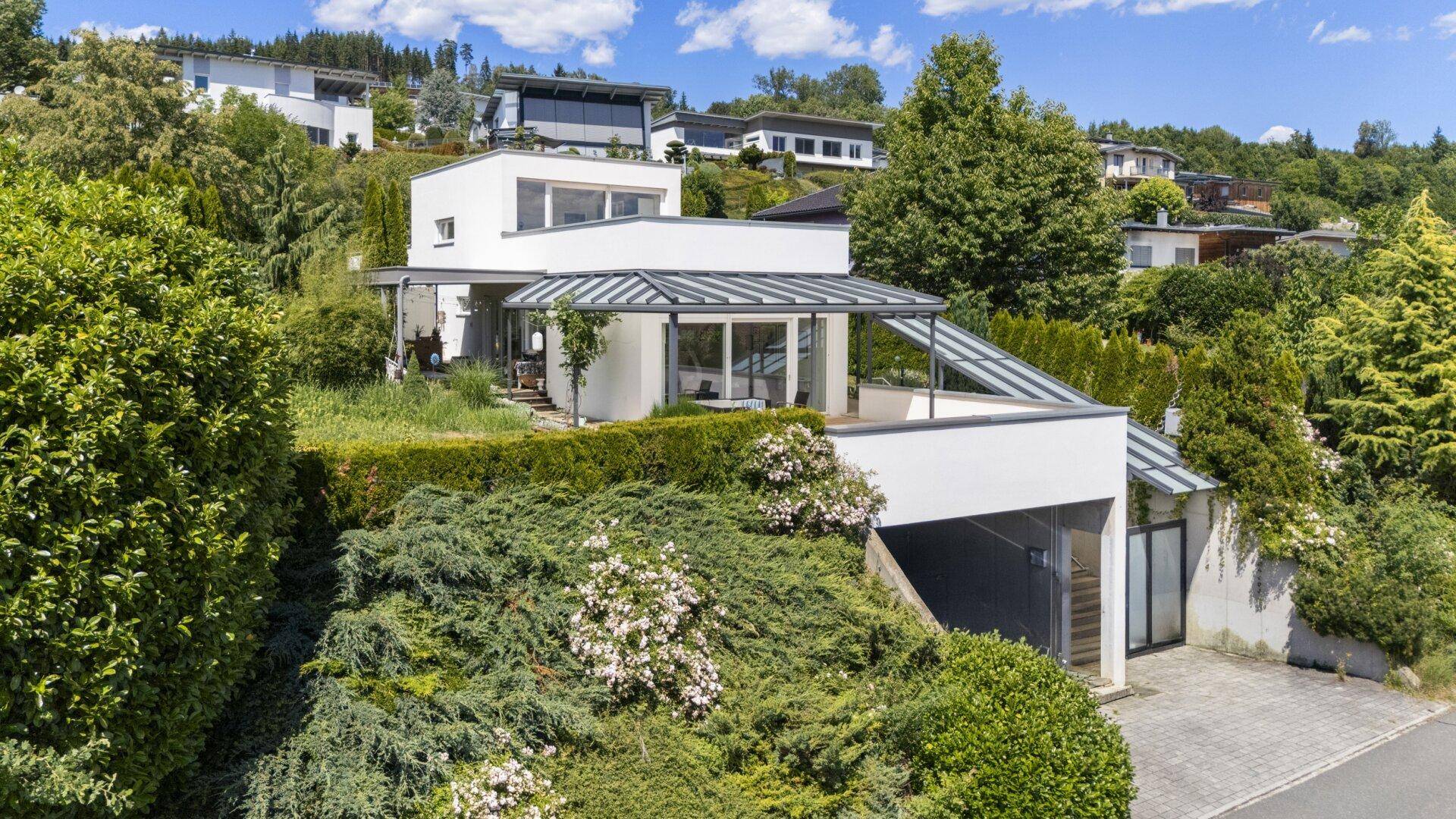 Panoramablick auf das moderne Haus mit Garage und Terrassenbereich, eingebettet in die grüne Hügellandschaft.