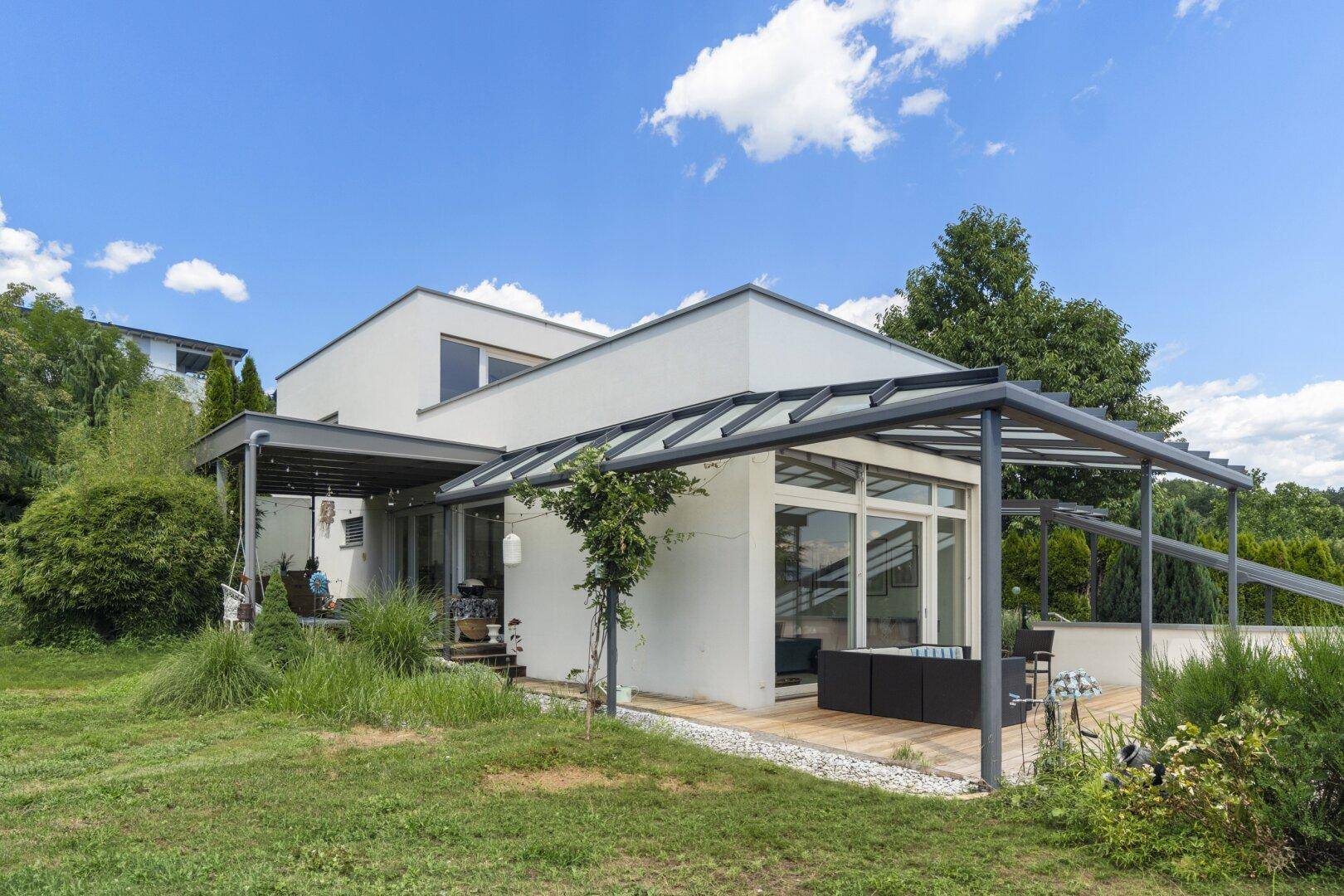 Ansicht des modernen Hauses mit großer überdachter Holzterrasse und Gartenbereich unter blauem Himmel.
