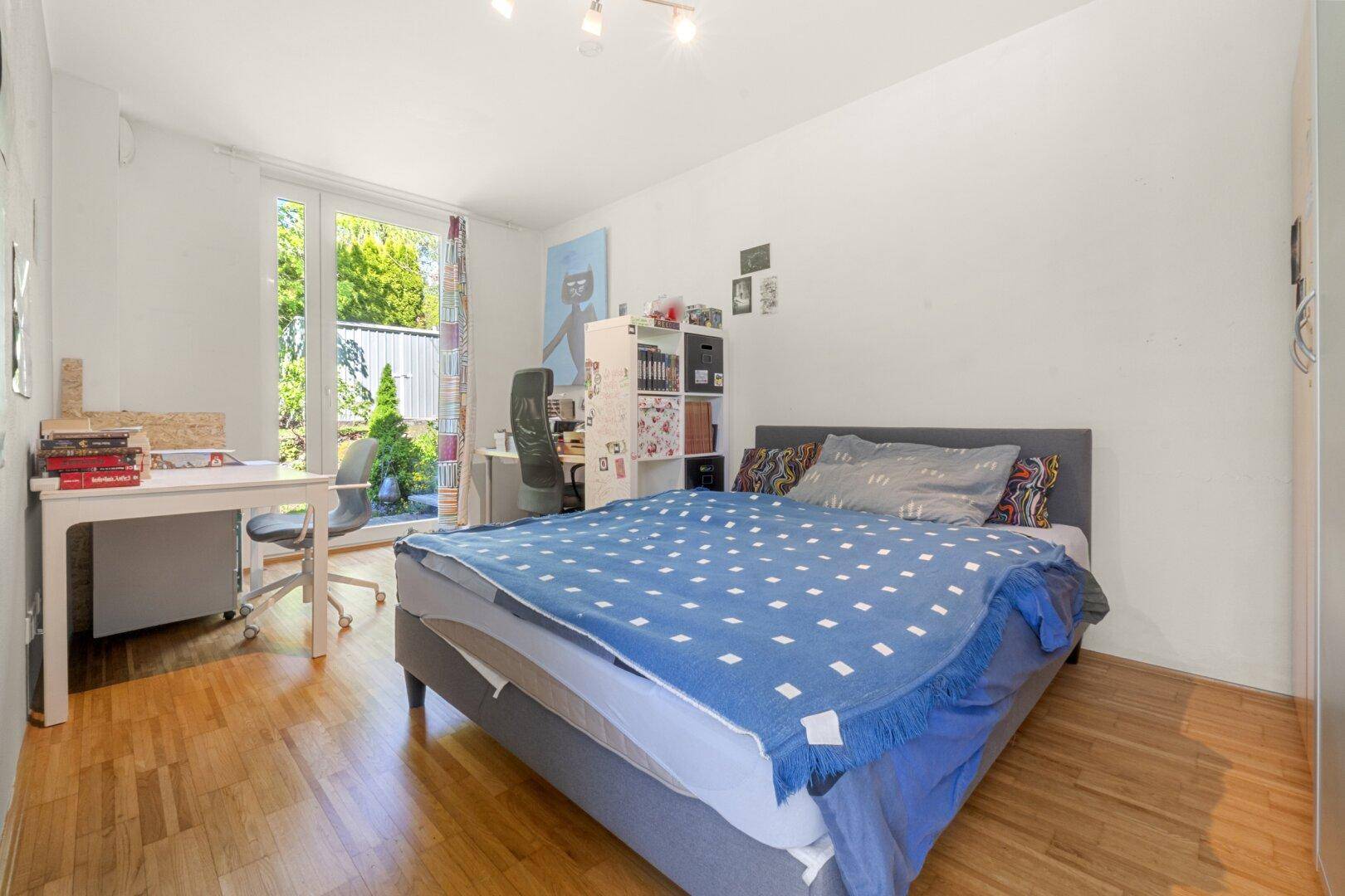 Freundliches Schlafzimmer mit Holzboden, Schreibtisch und Blick ins Grüne durch die Terrassentür.