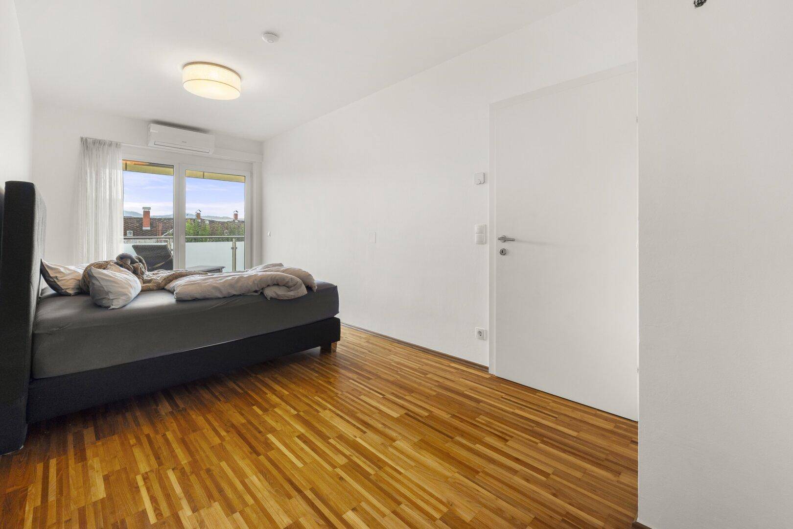 Helles Schlafzimmer mit Parkettboden, Klimaanlage und Zugang zu einem kleinen Balkon.