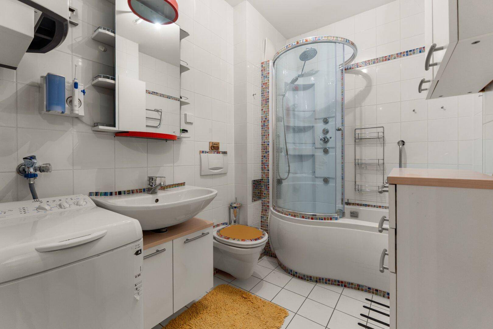 Helles Badezimmer mit weißen Fliesen, Duschkabine, Waschbecken und integrierter Waschmaschine.