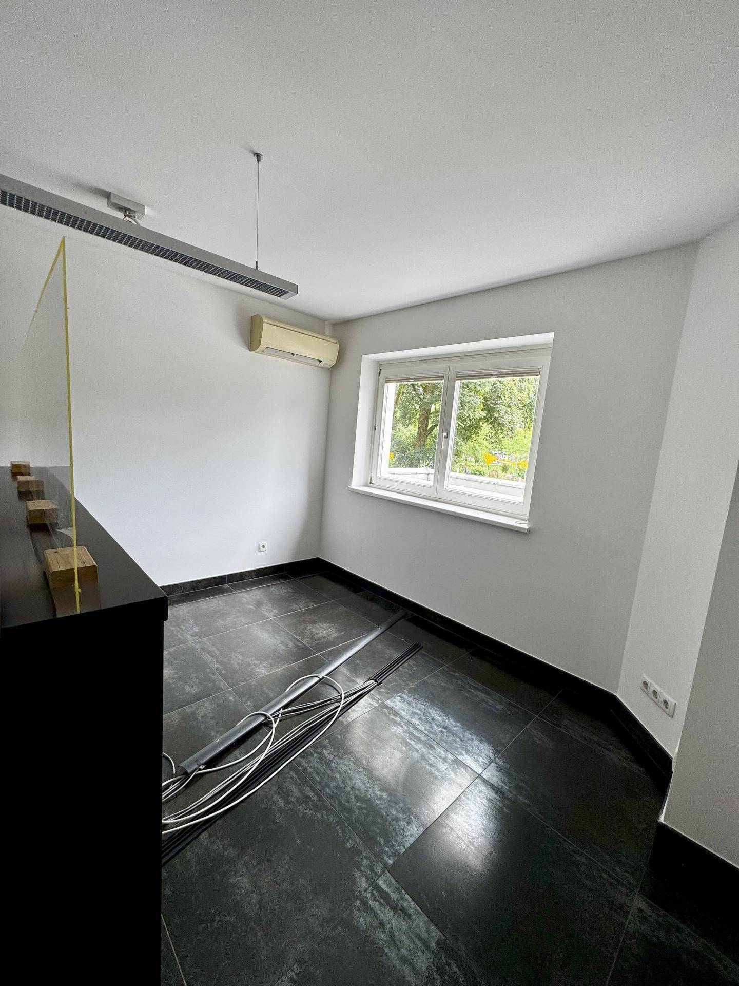 Helles Büro mit dunklen Fliesen, Klimaanlage und Fenster mit Blick ins Grüne.