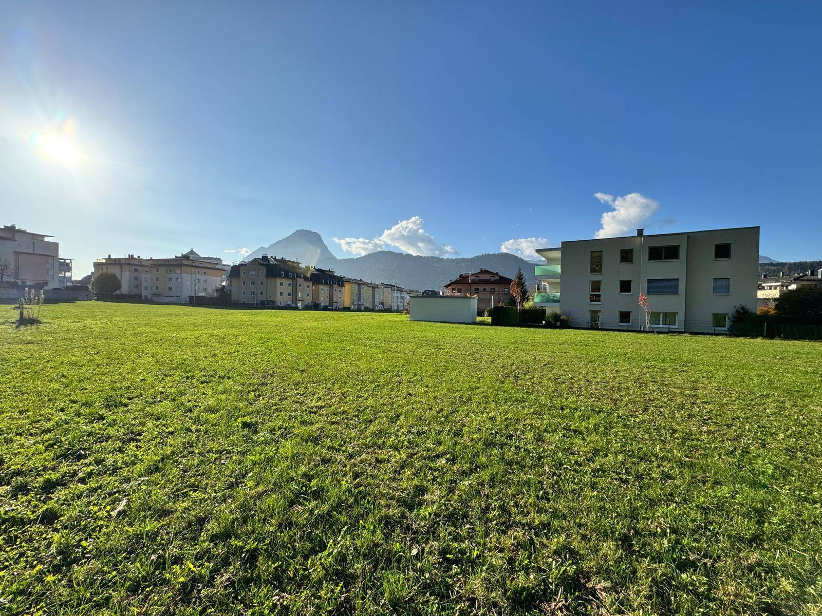 Große grüne Fläche mit städtischen Gebäuden und majestätischen Bergen unter einem klaren, sonnigen Himmel.