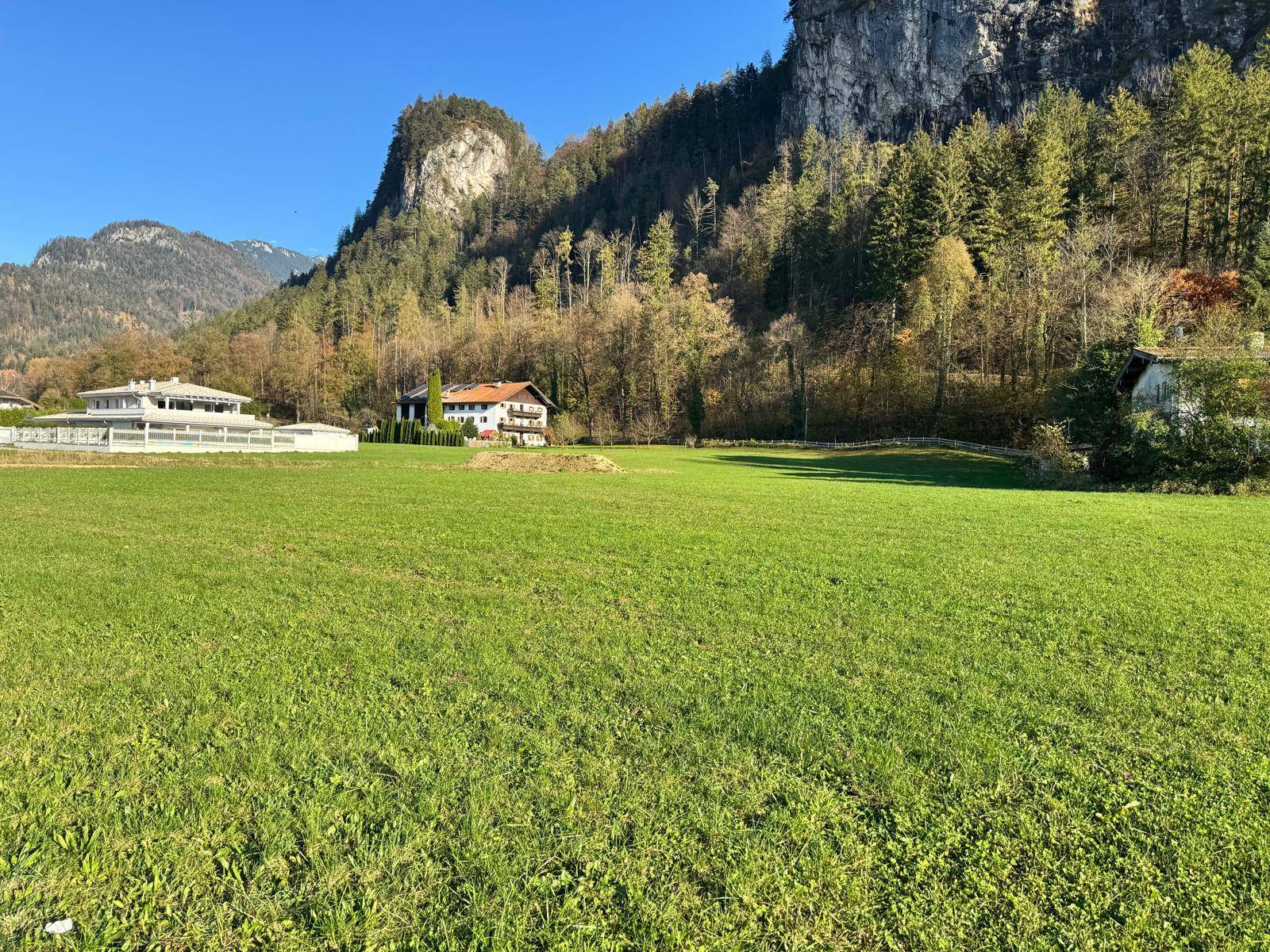 Ausgedehnte grüne Wiese mit einer Kulisse aus bewaldeten Bergen und vereinzelten Häusern in der Ferne.